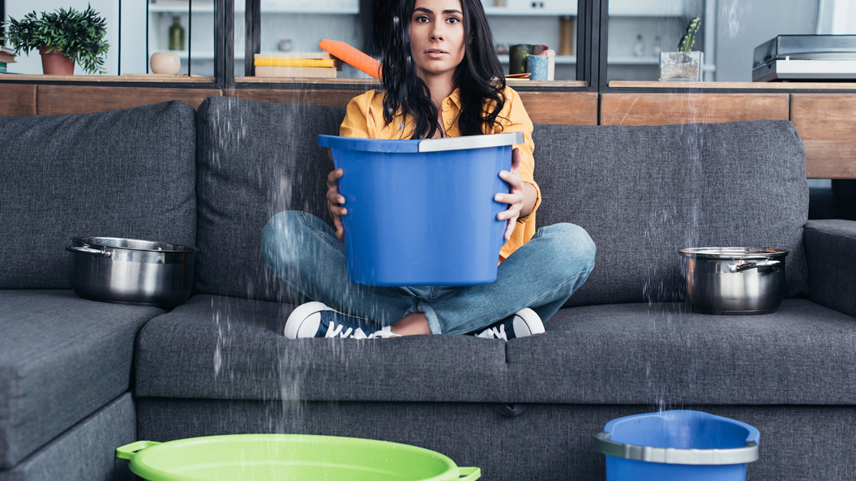 Brunette woman holding bucket on sofa during water damage in living room
