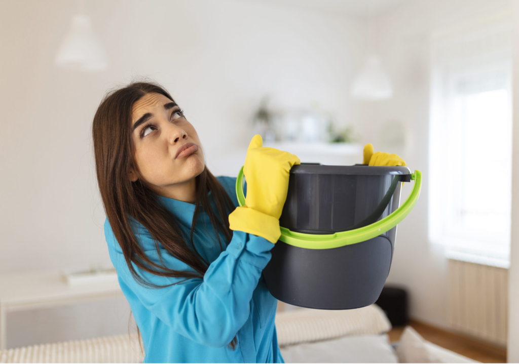 Shocked Woman Looks at the Ceiling While Collecting Water Which Leaks in the Living Room at Home. Worried Woman Holding Bucket While Water Droplets Leak From Ceiling in Living Room