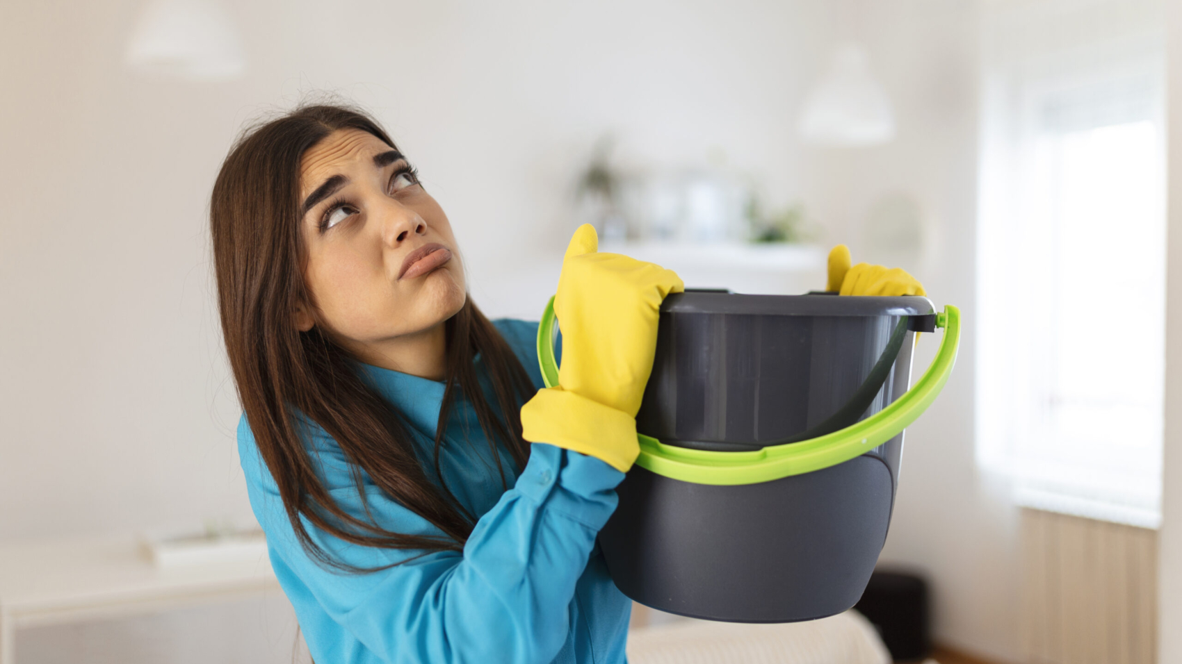 Shocked Woman Looks at the Ceiling While Collecting Water Which Leaks in the Living Room at Home. Worried Woman Holding Bucket While Water Droplets Leak From Ceiling in Living Room