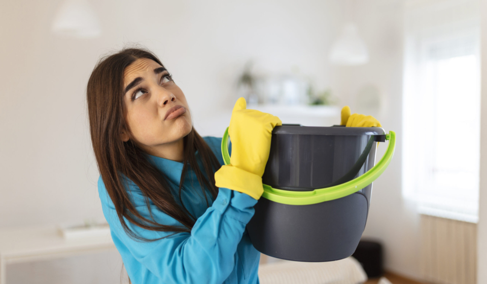 Shocked Woman Looks at the Ceiling While Collecting Water Which Leaks in the Living Room at Home. Worried Woman Holding Bucket While Water Droplets Leak From Ceiling in Living Room