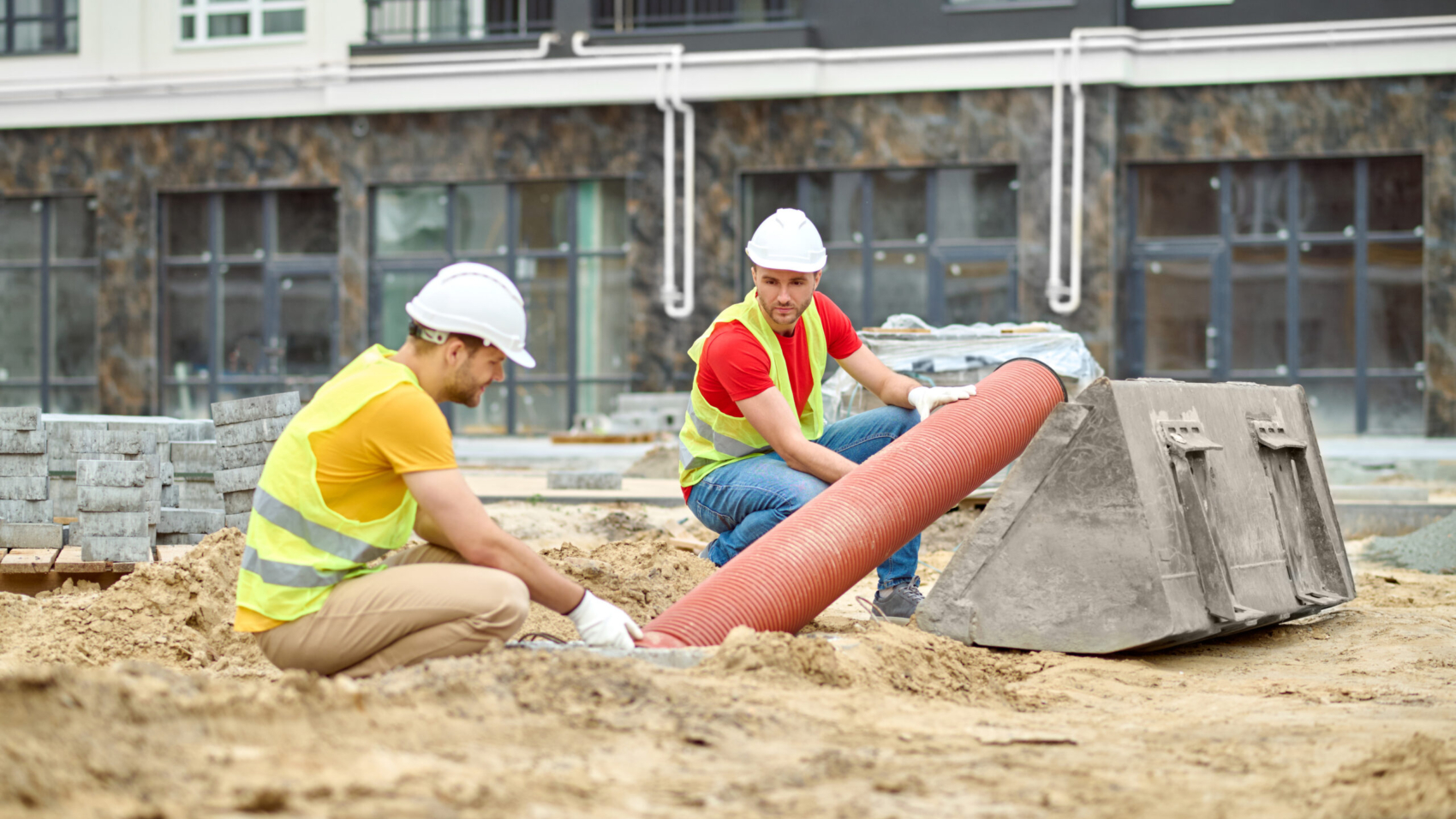 Working mood. Two men in protective helmet and bright vest crouched touching pipe on sand at construction site during day