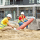 Working mood. Two men in protective helmet and bright vest crouched touching pipe on sand at construction site during day