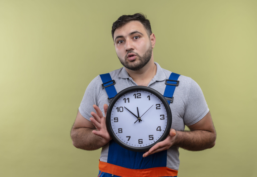 young builder man in construction uniform holding wall clock looking surprised standing over green background