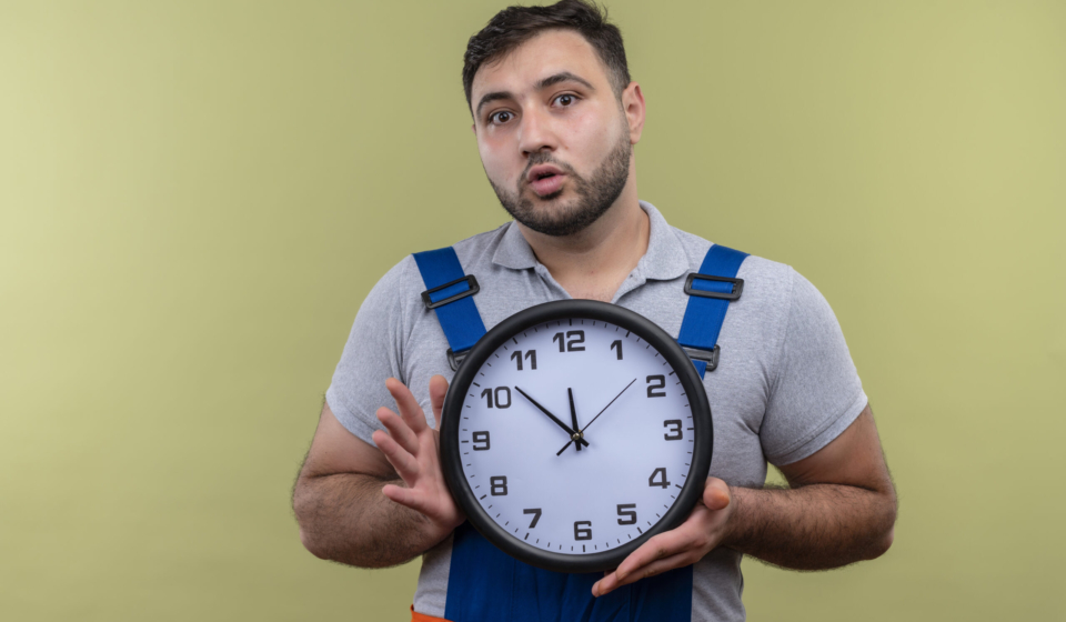 young builder man in construction uniform holding wall clock looking surprised standing over green background